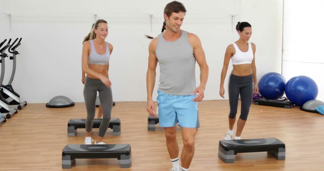 Three fit individuals are participating in a group step aerobics class with a trainer leading the exercise. Two women and one man are engaged in the workout, wearing sportswear. The room is bright and equipped with fitness equipment including steps and fitness balls. Perfect for promoting fitness classes, gym memberships, aerobic exercises, and healthy lifestyle.