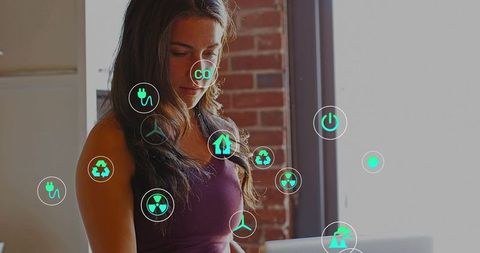 Standing woman viewing laptop with augmented green energy icons in modern office workspace