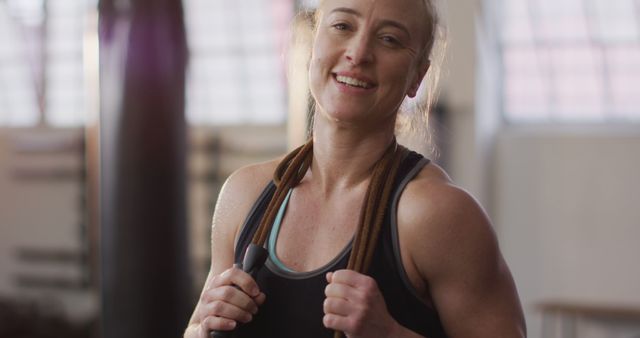 Woman holding skipping rope and smiling after exercising in gym. Could be used for advertisements or promotions related to fitness, health, athletic wear, and empowerment campaigns.