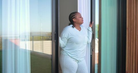 African american woman standing at sliding door enjoying sunlit balcony wearing loungewear