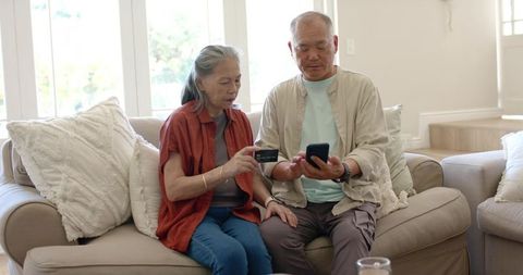 Asian senior couple tapping smartphone with credit card for mobile payment at home