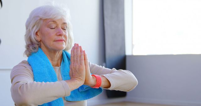 A senior Caucasian woman is practicing yoga with her eyes closed, hands pressed together in a prayer position, with copy space. Her peaceful expression and the towel around her neck suggest a focus on health and wellness.