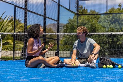 Couple sitting cross-legged and talking on blue padel court during sunny outdoor break