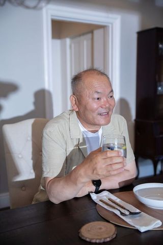 Asian senior man smiling while holding glass with lemon at cozy wooden dining table