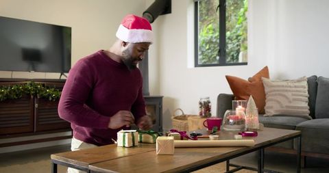 African american man wearing santa hat tying christmas gifts on living room table