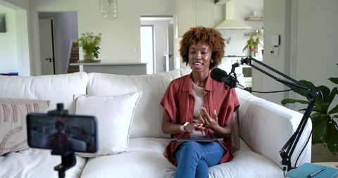 African american creator podcasting on sofa with microphone, tablet and smartphone