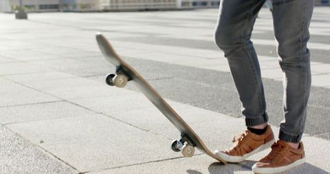 Balancing skateboard nose on sunlit plaza in cuffed gray jeans and brown sneakers