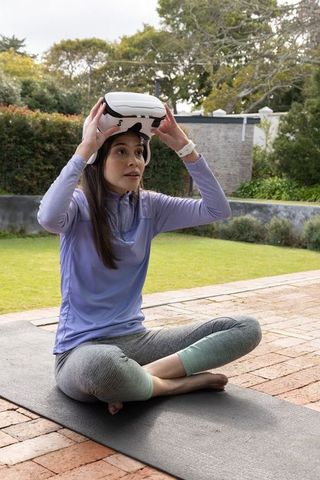 Young woman adjusting vr headset while sitting cross-legged on yoga mat in backyard garden