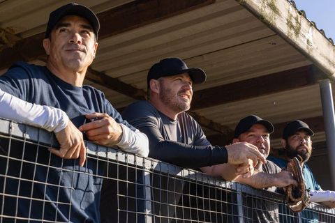 Diverse baseball teammates leaning on dugout fence watching game in sunlight