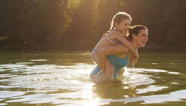 Mother carrying daughter piggyback in lake at golden hour smiling and playing in water
