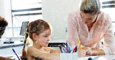 Young girl writing while teacher guiding in bright STEM classroom for elementary learning