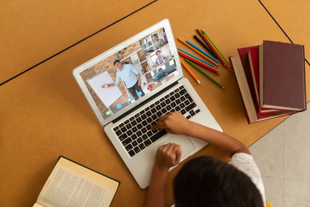 Student using laptop for attending virtual class while seated at table with open books and various colored pencils. This image is excellent for illustrating concepts such as distance education, e-learning, homeschooling, studying at home, technology in education, and remote teaching methods.