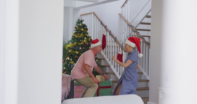 Senior couple is joyfully dancing in a beautifully decorated living room with a Christmas tree and gifts nearby. Both wearing Santa hats, are in the holiday spirit. Suitable for holiday greeting cards, advertisements focused on family celebrations, and promotional material for festive occasions.