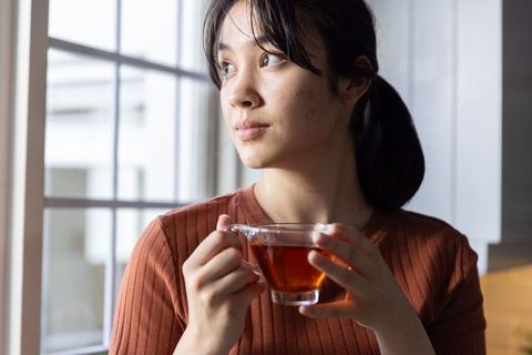 Asian woman holding glass cup of tea by window contemplating morning calm in cozy kitchen