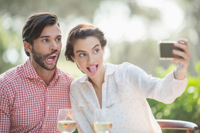 Young couple making funny faces while taking a selfie at an outdoor restaurant. They are enjoying their time together with wine glasses on the table. Perfect for use in advertisements, social media posts, blogs about relationships, leisure activities, or dining experiences.