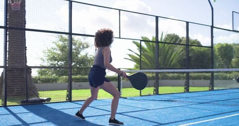 African american woman playing padel on blue outdoor court swinging paddle in action