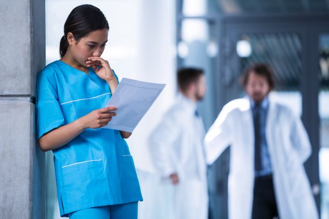 Nurse in blue scrubs thoughtfully examining a medical report in a hospital corridor. Two doctors are conversing in the background. Ideal for use in healthcare, medical, and hospital-related content, emphasizing patient care, medical examination, and teamwork in clinical settings.