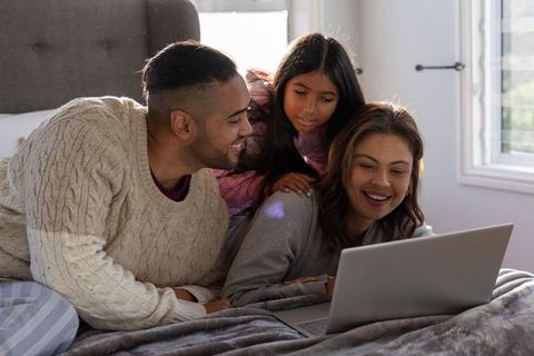 Smiling family of three watching laptop together on cozy sunlit bed, weekend home bonding