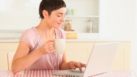 Woman Drinking Coffee While Using Laptop in Kitchen