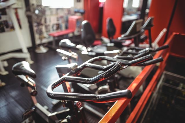 Close-up view of an exercise bicycle in a fitness studio. The image captures the handlebars and part of the bike, with other gym equipment visible in the background. Ideal for use in articles or advertisements related to fitness, health, gym equipment, and workout routines.