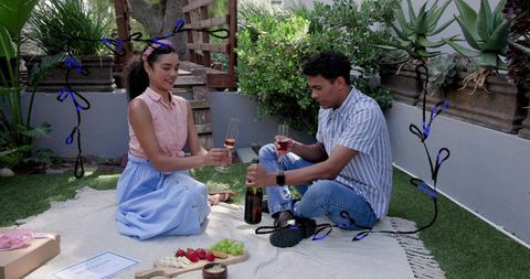 Couple toasting with wine on backyard picnic blanket with charcuterie and fresh fruit