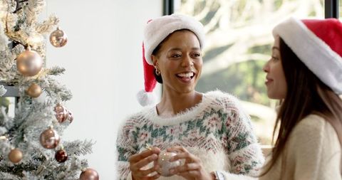 Diverse friends decorating christmas tree wearing santa hats and cozy festive sweaters