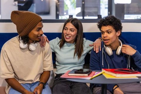 Multiracial teen friends chatting and studying on campus lounge sofa with notebooks