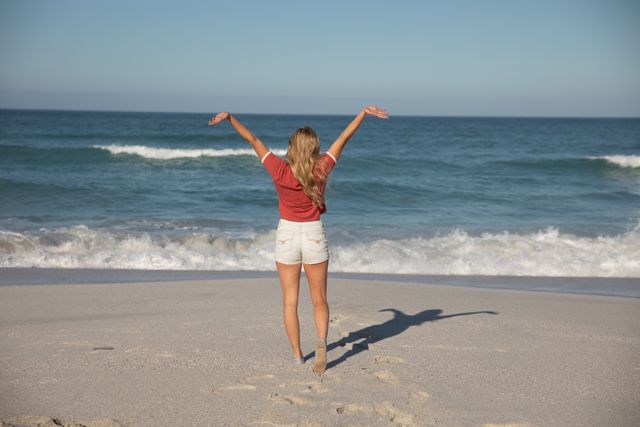 This image depicts a Caucasian woman standing on a sandy beach with her arms raised, facing the ocean on a sunny day. Ideal for use in travel brochures, summer vacation advertisements, wellness and relaxation promotions, and lifestyle blogs.