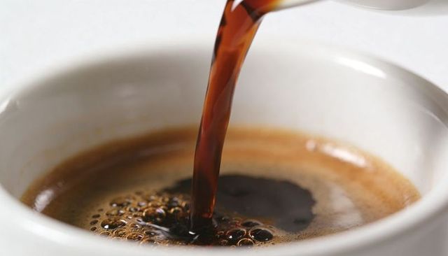 Pouring dark espresso stream creating crema and bubbles in white ceramic cup macro closeup