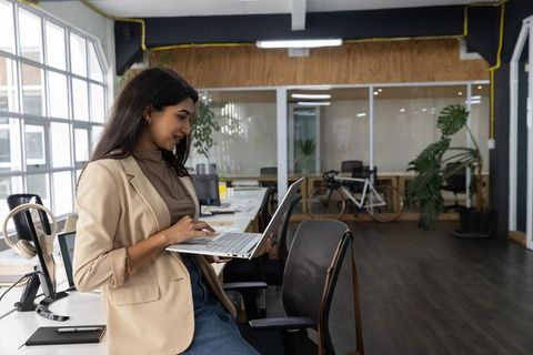 Indian professional leaning on desk working on laptop in modern open-plan coworking office
