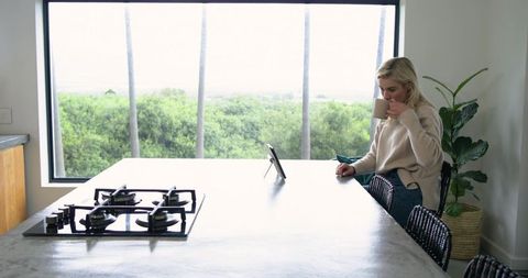 Blonde woman sipping coffee on wicker stool at minimalist kitchen island with window view