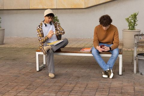Diverse male students sitting on bench checking watch and using smartphone on urban campus