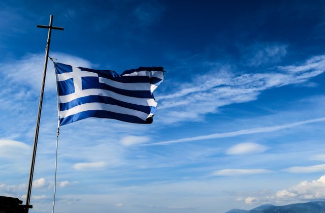 Greek flag waving proudly next to cross against clear blue sky, conveying strong sense of national pride and Greek heritage. Good for travel blogs, educational content on Greece, and cultural articles.