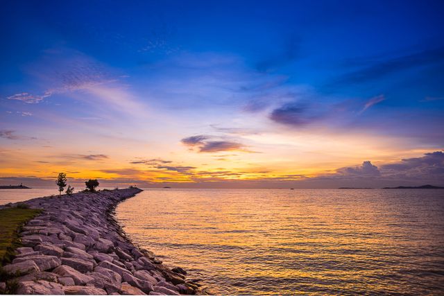A soothing scene of a sunset with an orange glow over a tranquil ocean, the horizon adorned with dramatic skies and clouds. The rock jetty stretches out into the calm waters, creating a picturesque view ideal for meditation, backgrounds, design inspirations, travel promotions, and calming visuals in media.