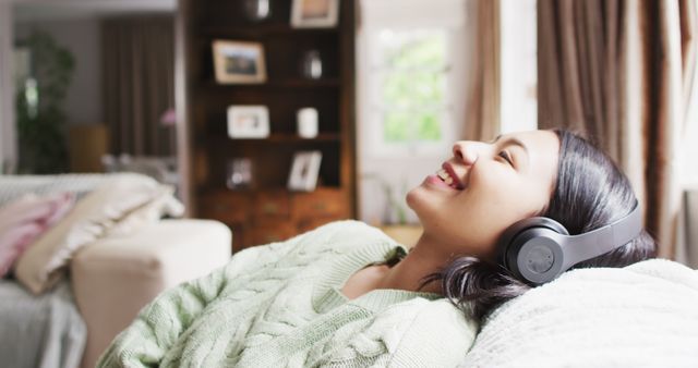 Young woman lying on a sofa wearing headphones, immersed in music in a cozy living room. Ideal for lifestyle, home, and leisure concepts. This can be used for advertisements for headphones, music streaming services, or relaxing home atmospheres.