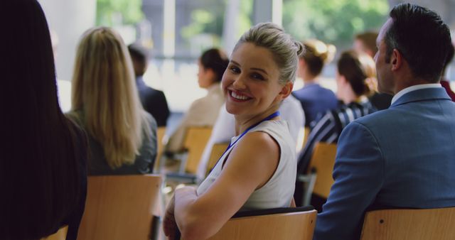 A blonde woman in formal attire, with a name tag, smiling warmly while seated among other professionals at a business conference. Ideal for themes related to networking, professional development, corporate events, seminars, or business meetings.