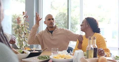 Diverse couple laughing and sharing salad at sunny family lunch table, cozy gathering