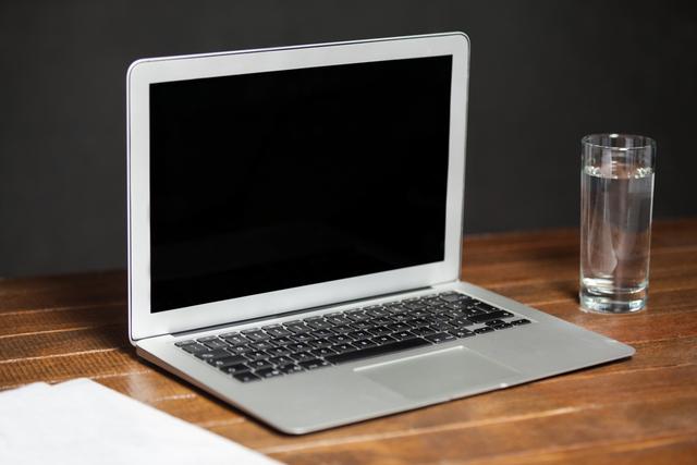Open laptop sits on a wooden table with a glass of water beside it. This image is ideal for use in articles about modern workspaces, home offices, and the importance of staying hydrated while working. It can also be used for technology-related advertisements, blog posts, or website backgrounds.