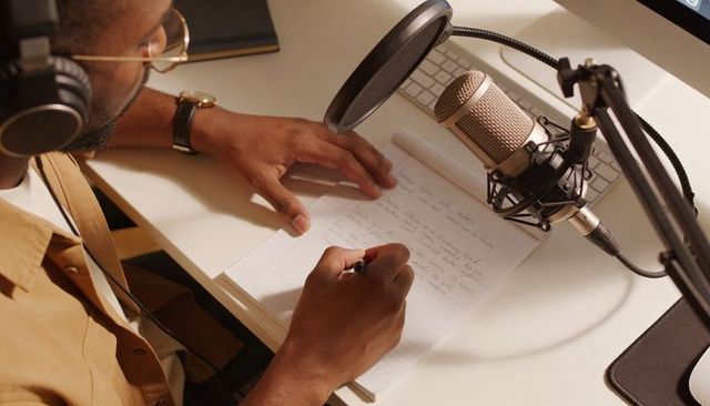 Recording podcaster writing notes at home desk with condenser mic and headphones