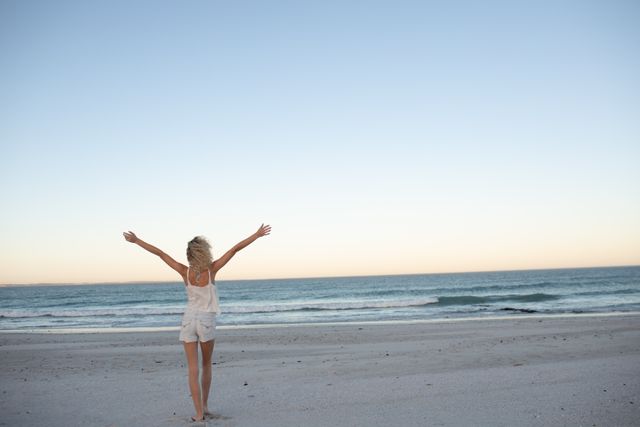 Woman standing on sandy beach with arms outstretched towards ocean at sunset. Ideal for use in travel brochures, wellness blogs, lifestyle magazines, and advertisements promoting relaxation, freedom, and beach vacations.