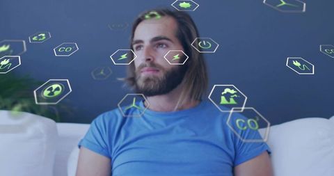 Gazing bearded man sitting on white sofa wearing blue shirt amid floating eco icons