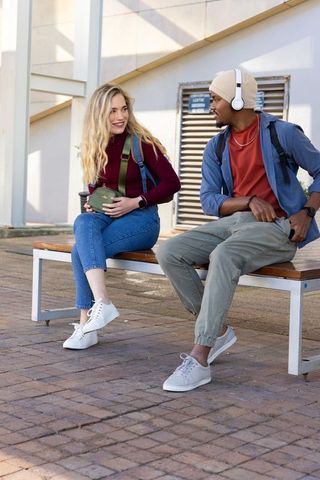 Young diverse friends sitting and talking on urban bench with backpacks and headphones