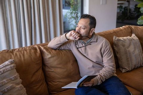 Asian man sitting on leather sofa holding book and glasses, contemplative daylight portrait
