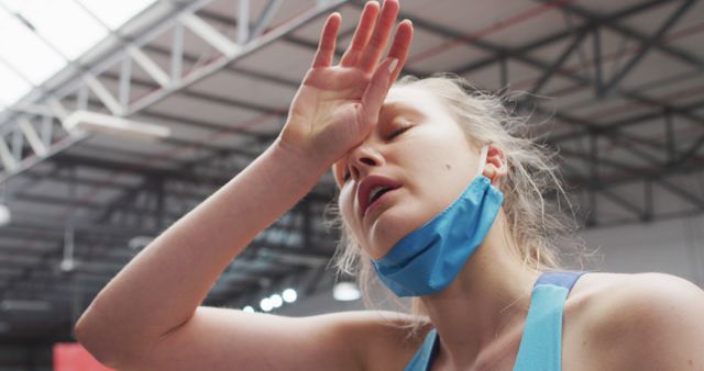 Caucasian woman wearing lowered face mask wiping brow at gym. blonde woman taking a break from her workout, wiping sweat from her brow with hand. hygiene at gym during coronavirus covid 19 pandemic