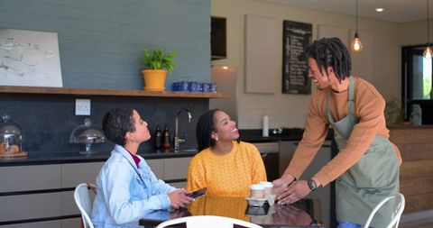 African-american barista serving coffee to smiling diverse women at cozy modern cafe table