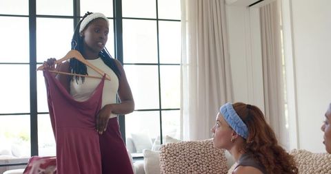 African american woman holding burgundy dress while friends comparing outfits