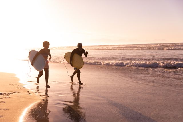 Couple running on beach with surfboards at sunset, enjoying a carefree moment. Ideal for themes of love, togetherness, retirement, and holiday enjoyment. Perfect for travel brochures, lifestyle blogs, and advertisements promoting beach vacations and aquatic sports.