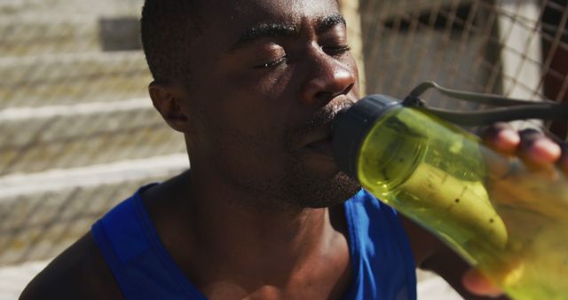 Tired african american man sitting and drinking from water bottle, taking break in exercise outdoors. fitness, healthy and active lifestyle concept.