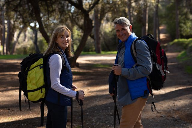 Portrait of a happy senior Caucasian couple enjoying time in nature together, hiking in forest on a sunny day, holding Nordic walking sticks smiling to camera.