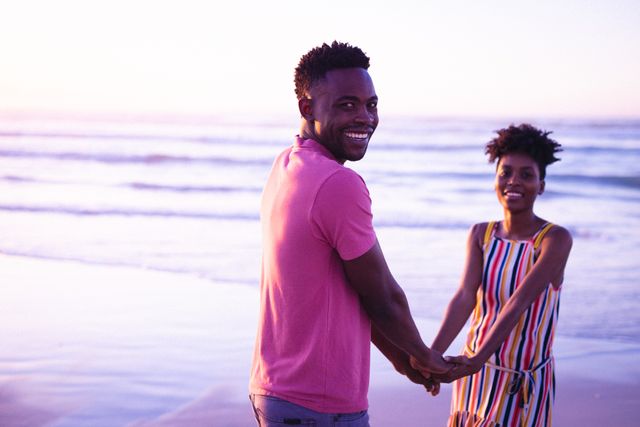 This image captures a joyful African American couple holding hands at the beach during sunset. The man is smiling at the camera while the woman stands behind him, both enjoying a romantic moment by the sea. Perfect for use in advertisements, travel brochures, relationship blogs, and lifestyle magazines to convey themes of love, happiness, and vacation.